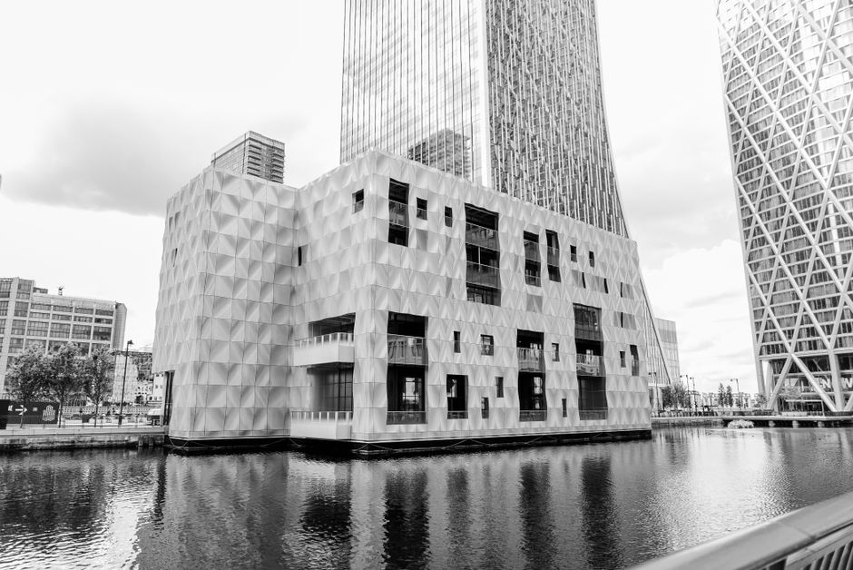 A modern, multi-story building with a distinctive geometric facade featuring a pattern of diamond-shaped panels, situated beside a waterway in Canary Wharf. The building's exterior is composed of slee