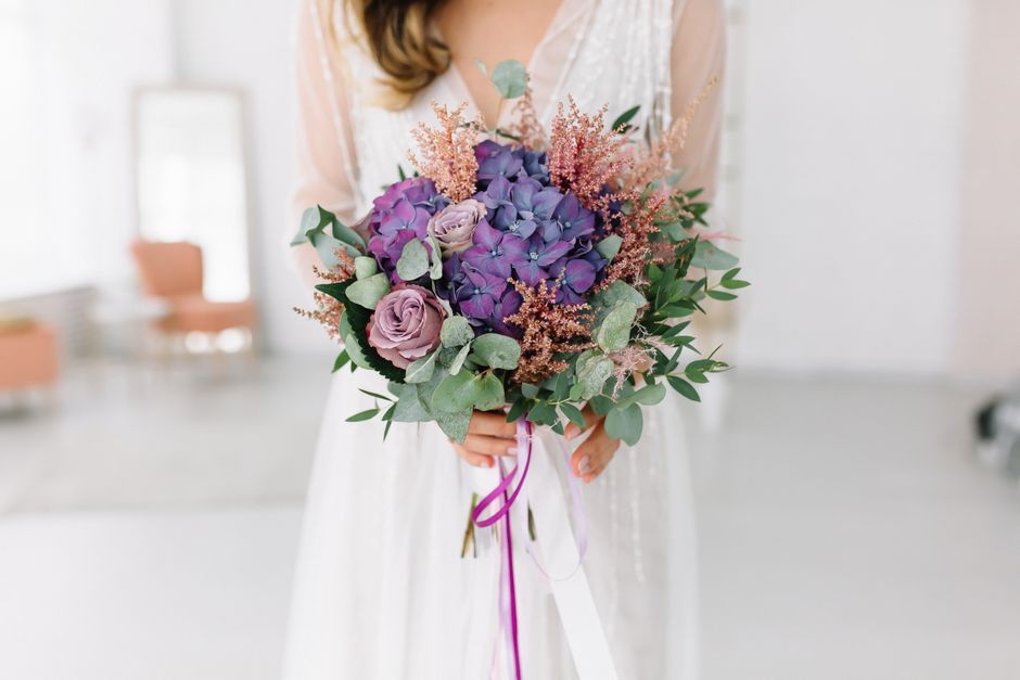 A bridal bouquet featuring a combination of deep purple hydrangeas, soft lavender roses, and wispy pink astilbe, accented with muted green eucalyptus leaves. The arrangement is held by a woman dressed
