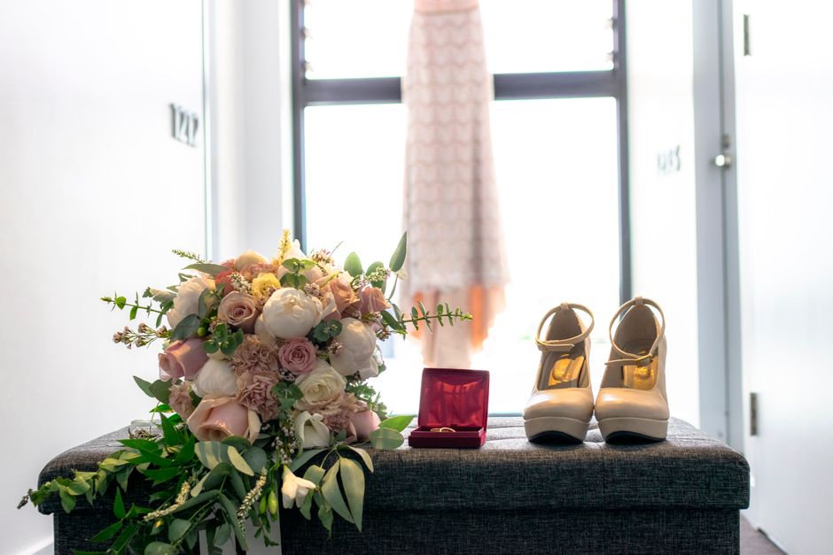 A floral arrangement of pastel pink and white roses, peonies, and greenery is displayed on a dark fabric bench near a bright window, with a pair of white bridal shoes with gold accents placed beside i