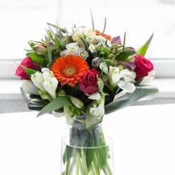 Brimming with modern elegance, this sophisticated bouquet artfully captures the contemporary Docklands spirit, blending bold blooms and nuanced textures to stunning effect. Prominent gerbera daisies in fiery orange and soft peach immediately draw the eye, their textured petals and deep brown centers glistening in natural daylight-a reflection of the vibrant ambiance found around Canada Square. Interwoven between them, luxurious magenta roses rest with timeless poise, their tightly clustered petals suggesting both passion and grace. Scattered throughout, the white blossoms of alstroemeria and hints of pale pink petals brighten and soften the composition with airy lightness. The flowers are hugged by a cascade of glossy, curved aspidistra leaves cradling the base, creating a strong, rounded foundation evocative of Docklands' blend of lush parks and architectural grandeur. Further enriched by supple, pointed greenery, every detail is meticulously curated and anchored in a tall, clear glass vase-showcasing the freshness and premium quality appreciated by local residents. Whether presented at a riverside gathering, a corporate occasion, or as a heartfelt Docklands delivery, this arrangement embodies joy, gratitude, and the beauty of urban nature, making it a centerpiece that captures the soul of the area.